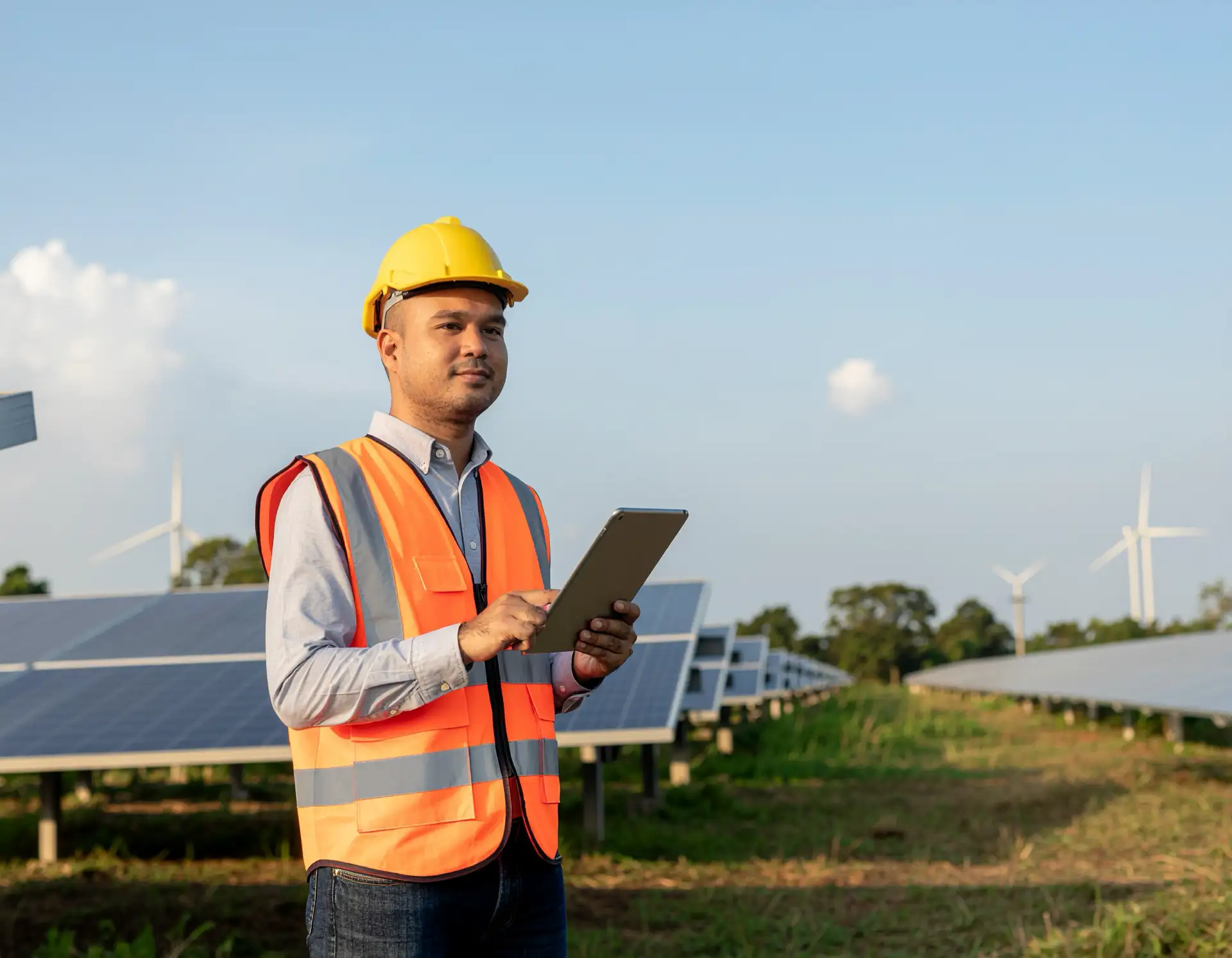 Man in yellow hard hat and orange vest holds a tablet in a solar panel field