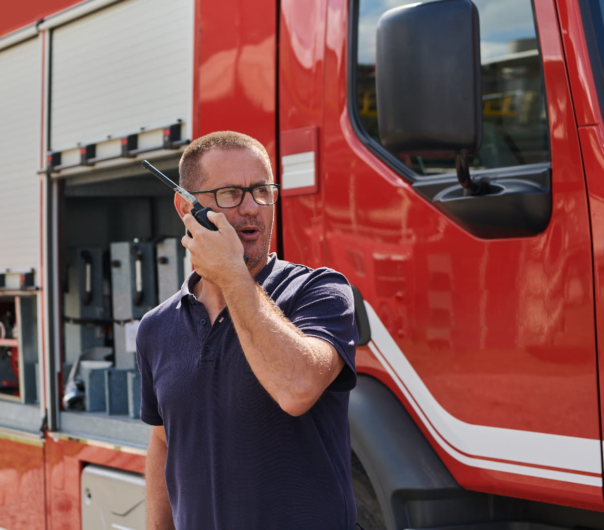 A firefighter stands before a modern firetruck talking into a device to help illustrate first responders IoT connectivity needs