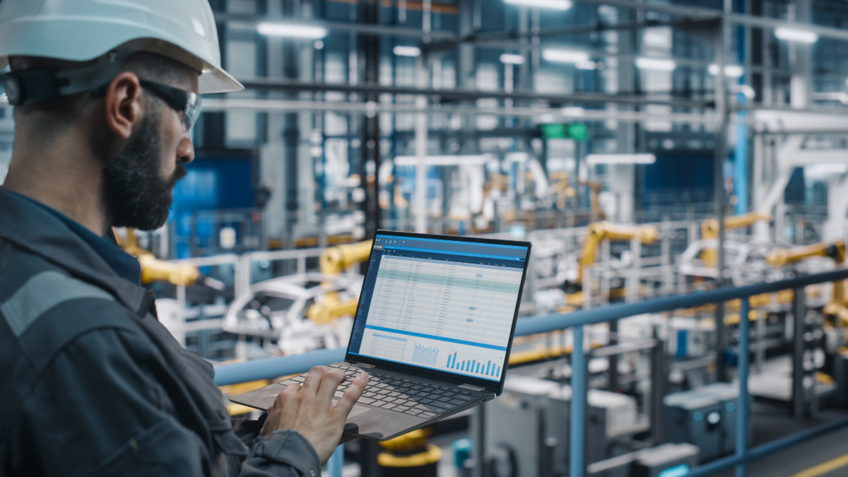 Car Factory Engineer in Work Uniform Using Laptop Computer to review IoT logistics connectivity at Automotive Industrial Manufacturing.