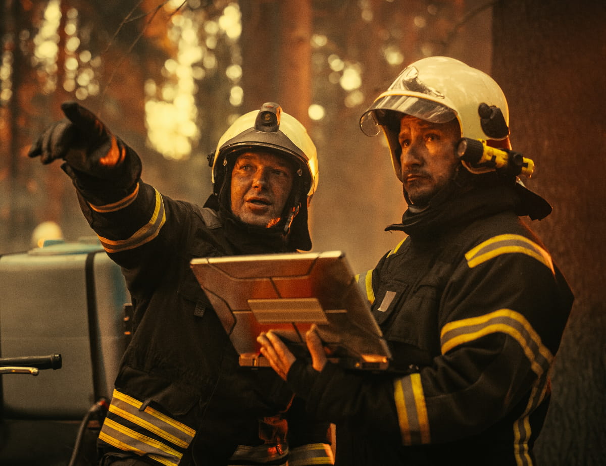 Portrait of Two Professional Firefighters Standing Next to an All-Terrain Vehicle, Using a Heavy-Duty Laptop Computer to illustrate first responder operations.