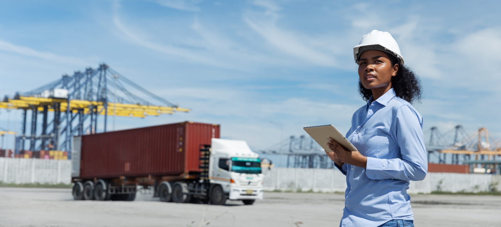 A female logistics officer in a hard hat observes cargo operations at a shipping port. Holding a tablet, she monitors container trucks and cranes involved in global transport and export enterprise IoT connectivity logistics operations.