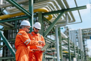 Oil and gas engineers in orange protective suits and hardhats look at a laptop with several pipelines behind them.