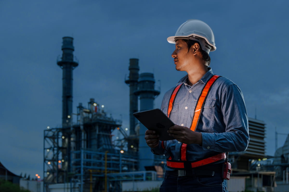 A worker looking at an oil operation in the background at dusk with the glowing screen of a tablet in his hands to help illustrate IoT for oil and gas